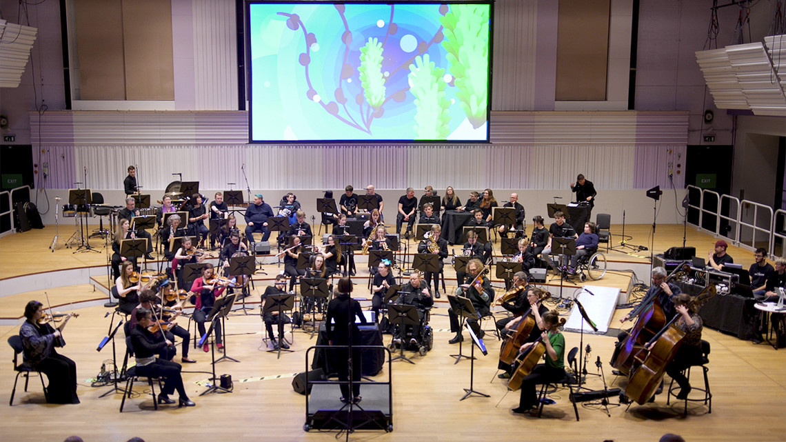 An orchestra of musicians on stage, some in wheelchairs, with a large screen behind projecting graphics.