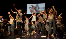 A group of performers on stage with arms outstretched in different directions. A screen behind showing a hand reaching out.