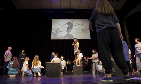 Performers on a stage, one woman in the centre stood on a box, a screen showing a video at the back of the stage