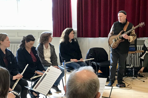 A guitar player talking to a group of musicians sat in a circle.