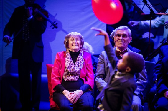 An older man and woman seated watching a young boy playing with a red balloon.
