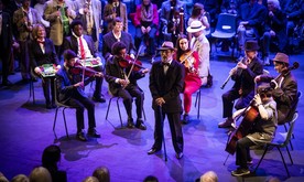 An older man standing in front of a group of instrumentalists on stage.