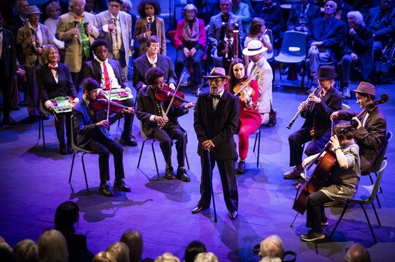 An older man standing in front of a group of instrumentalists on stage.