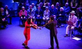 A young woman and young man playing violins on stage facing each other, seated rows of performers behind.
