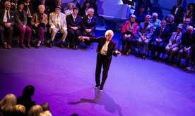 An older lady dancing on stage, seated rows of performers behind.