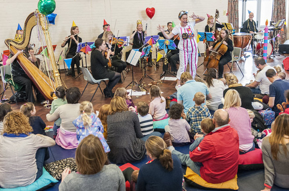 An audience of young children and adults watching a presenter and orchestra.