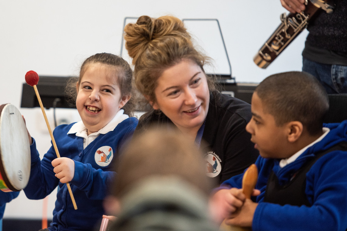 Two young children playing percussion instruments with a school teacher.