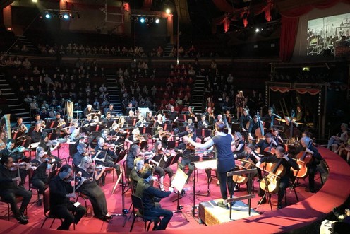A woman conducting a large orchestra and large childrens choir in a circular venue with a screen showing a film up above the orchestra.