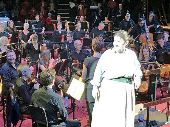 A woman in costume and a headset microphone in front of a large orchestra
