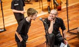 A young white boy talks into a microphone on stage with a white man in glasses crouched to the side looking at him