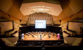 The view from the back of a concert hall of a small orchestra of musicians sat socially distanced on a stage with a screen projected above them.