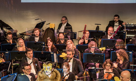 Two young producers dressed as a bee and lion present in front of an orchestra. A screen behind shows a musical score.