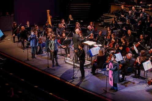 Young producers on stage in front of the orchestra talking into microphones.
