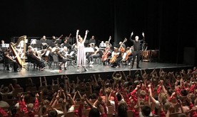 A presenter on stage in front of the orchestra waving to a large audience of children.